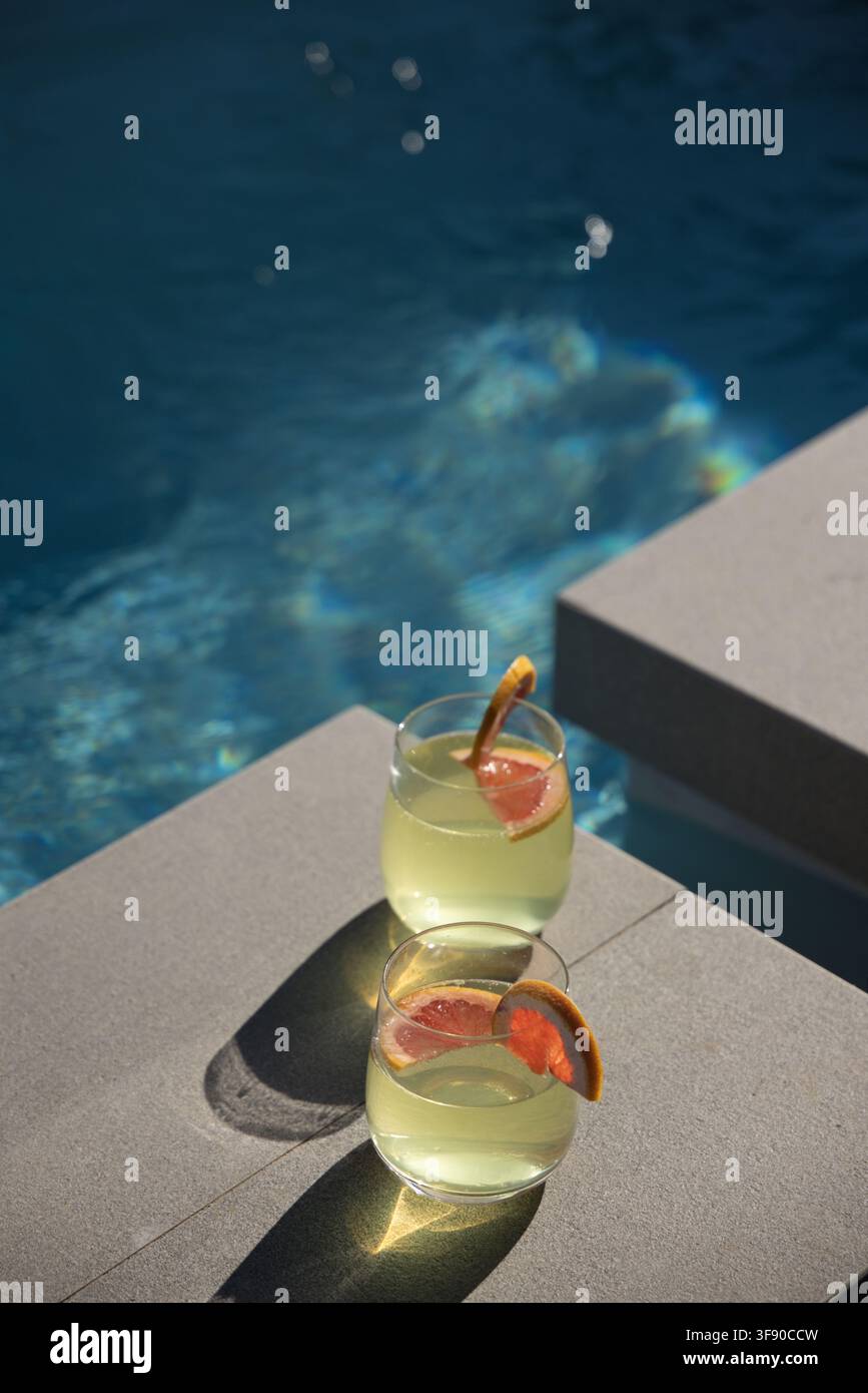 Zwei stiellose Gläser, die auf hellgrauen Poolstufen stehen, grünes Getränk, Zitrusfrüchte am Rand, Wasser glänzt Stockfoto
