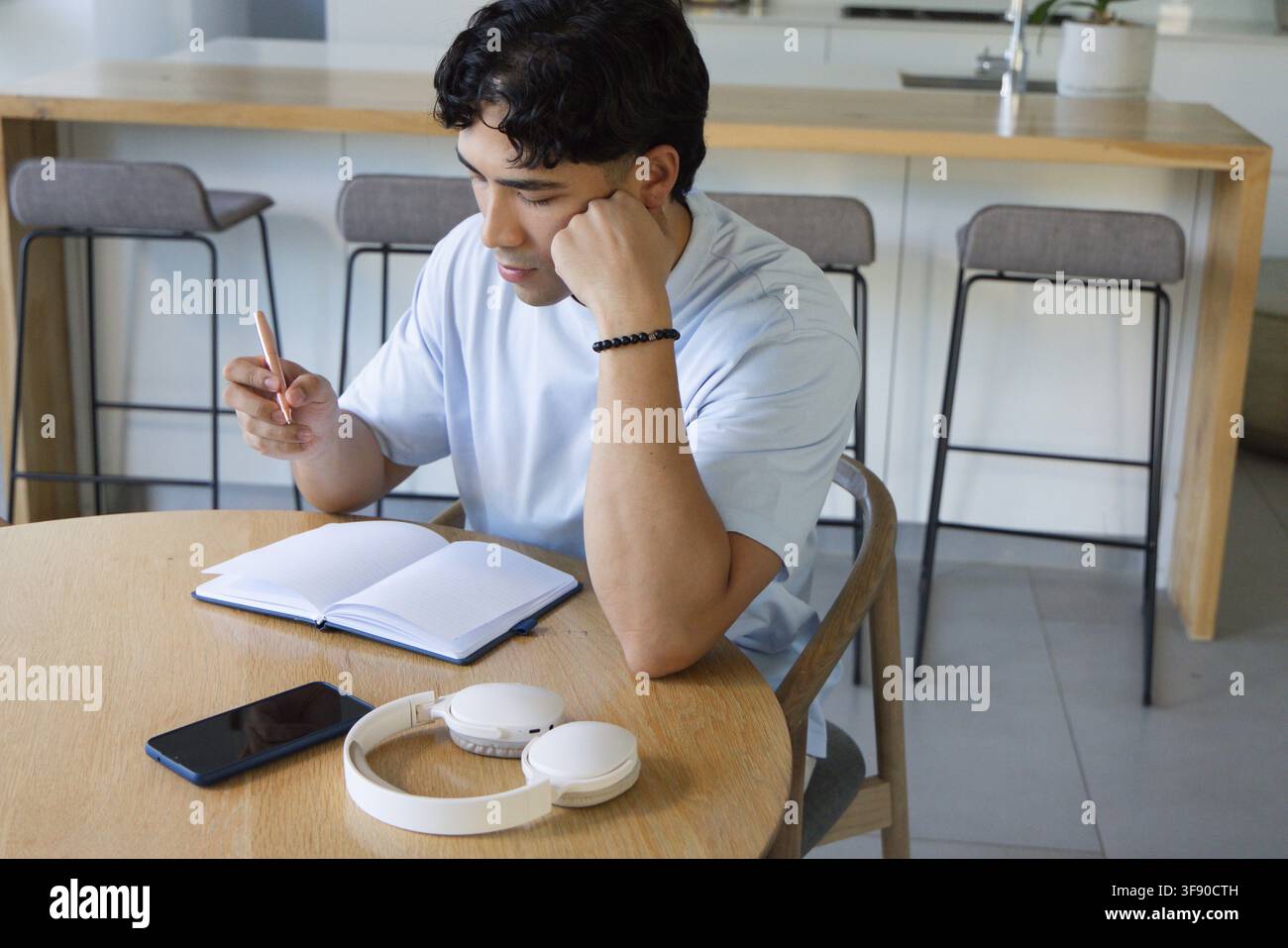 Asiatischer Mann Anfang 20 sitzend am Küchentisch und schreibt mit einem Stift in ein Notizbuch, Telefonkopfhörer Stockfoto