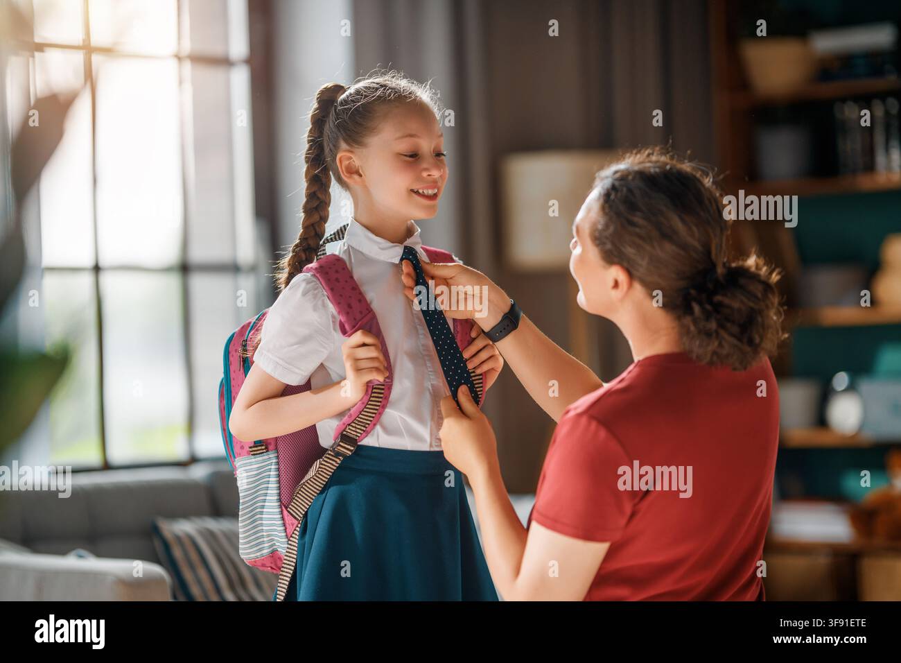 Warmer und freudiger Moment, in dem eine fürsorgliche Mutter ihrer lächelnden Tochter hilft, sich auf den Schultag vorzubereiten. Das Kind, das einen Rucksack und eine Uniform trägt, sieht aufgeregt aus Stockfoto