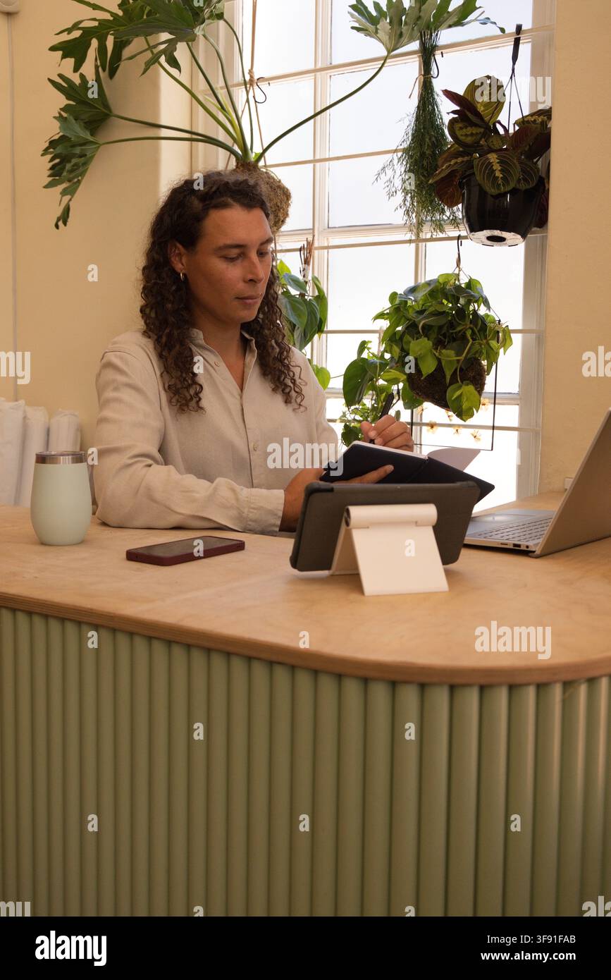 Nicht-binäre/r Afrikaner/in in beigem Hemd sitzt an geriffelter Theke und verwendet ein Tablet mit offenem Laptop daneben Stockfoto
