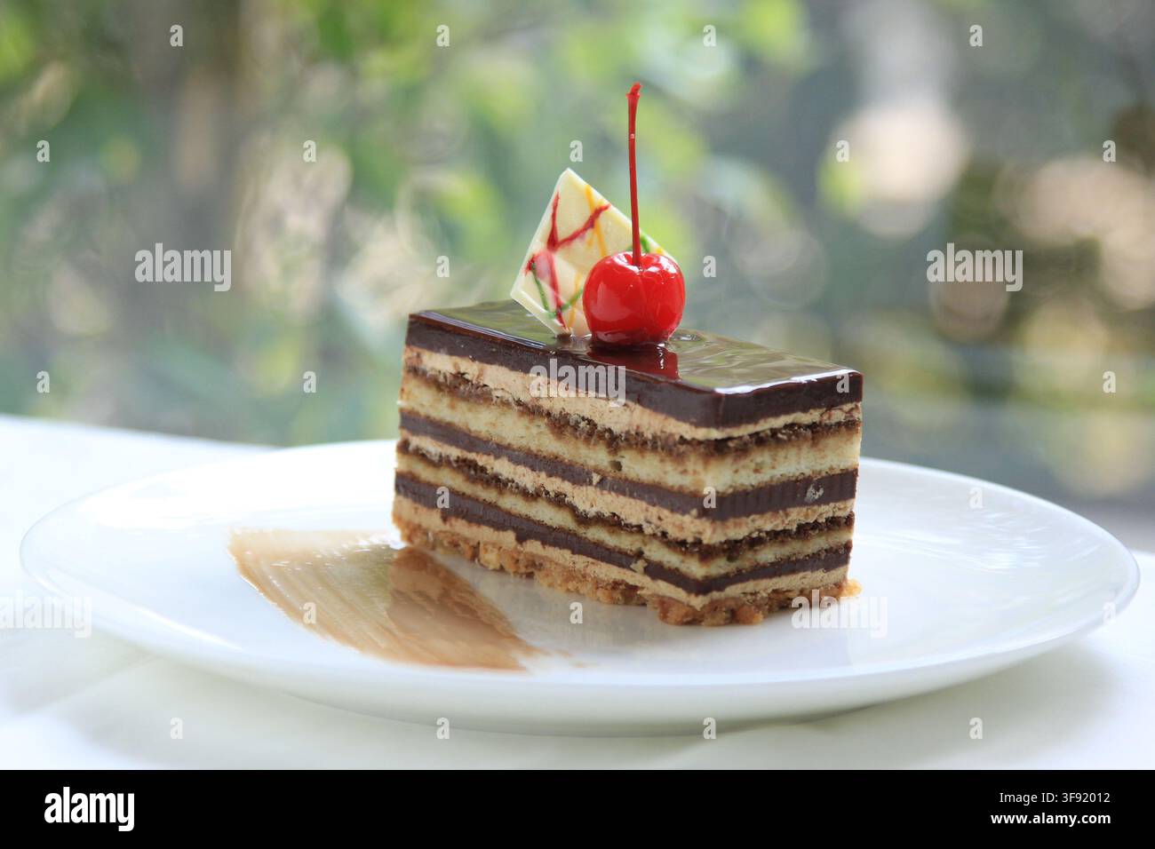 Nahaufnahme einer Scheibe geschichteten Schokoladenkuchens oder Opernkuchens, der mit Schokoladenglasur und einer Maraschino-Kirsche auf einem weißen Teller garniert ist. Stockfoto
