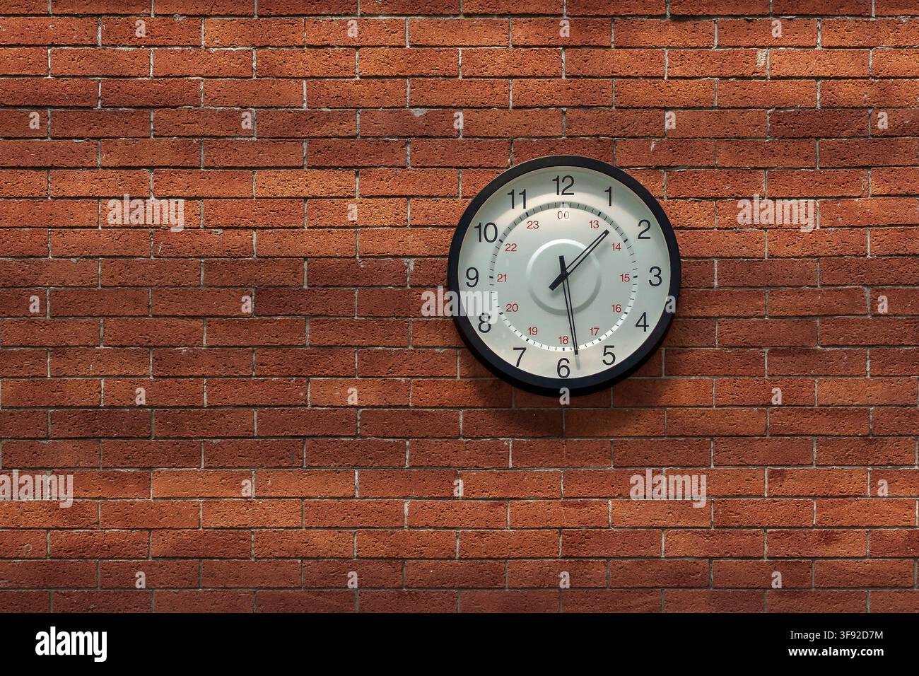 Eine klassische runde analoge Wanduhr mit schwarzem Rand und weißem Zifferblatt, zentriert auf einem strukturierten roten Ziegelwandhintergrund montiert. Stockfoto