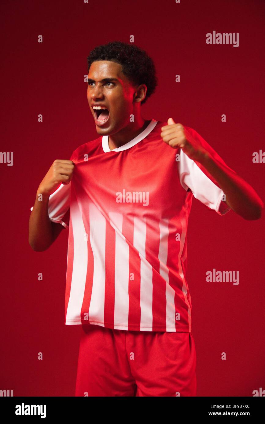 Afroamerikanischer Mann zieht rotes und weiß gestreiftes Trikot weg und schreit vor rotem Studiohintergrund Stockfoto