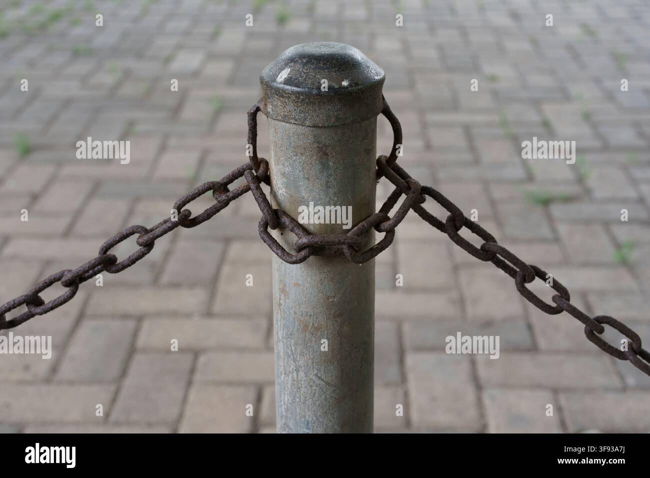 Nahaufnahme eines verwitterten Metallpfostens, der eine schwere rostige Eisenkette als Absperrung über Kopfsteinpflasterblöcken im Freien sichert Stockfoto