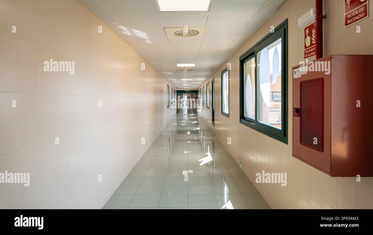 Langer steriler Flur in einer modernen medizinischen Einrichtung mit heller Beleuchtung, reflektierenden Fliesenböden und an der Wand montierter Brandschutzausrüstung in quie Stockfoto