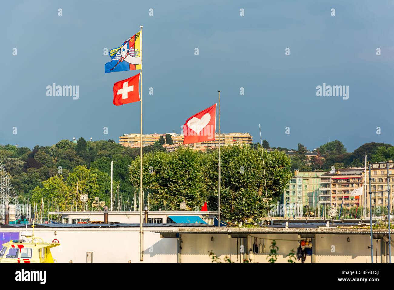 Schweizer und farbenfrohe Flaggen wehen über einem Yachthafen am Ufer von Genf, wobei Bäume, Wohnhäuser und ein blauer Himmel die städtische Kulisse bilden. Stockfoto