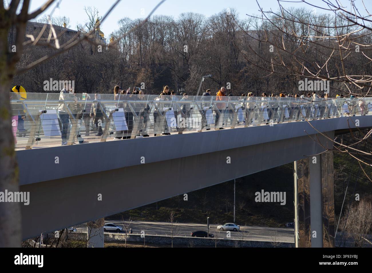 Kiew, Ukraine – 15. März 2026: Menschenmenge, die an einem klaren Tag auf einer malerischen Fußgängerbrücke mit Glasgeländern spaziert, und präsentiert Gemeinschafts-Lei Stockfoto