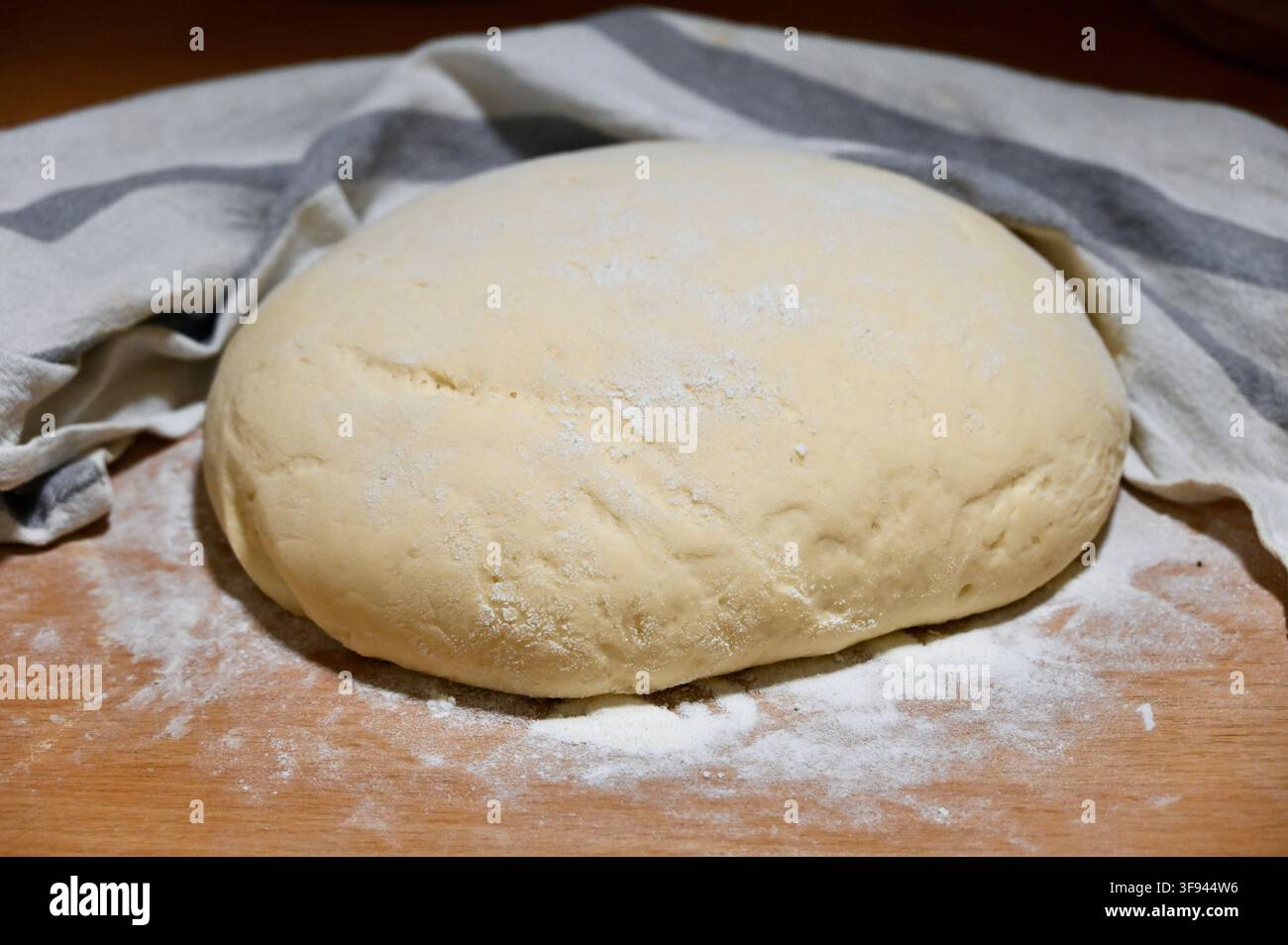 Kommerzielles Nahaufnahmefoto eines großen Klumpens weicher, ungebackener Hefebrodteig, der auf einer rustikalen Holzplatte liegt, die mit Mehl bestäubt ist, mit einem gestreiften Küchentuch im Hintergrund. Stockfoto