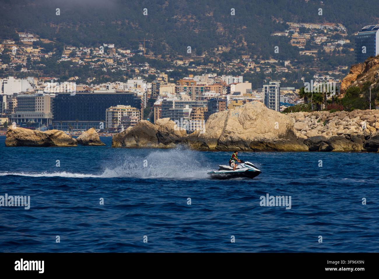 Calp, Spanien - 11. Juli 2027: Blick auf eine Person, die auf einem Jet-Ski auf dem Mittelmeer fährt, mit der Küstenstadt Calp und felsigen Klippen im Hintergrund Stockfoto