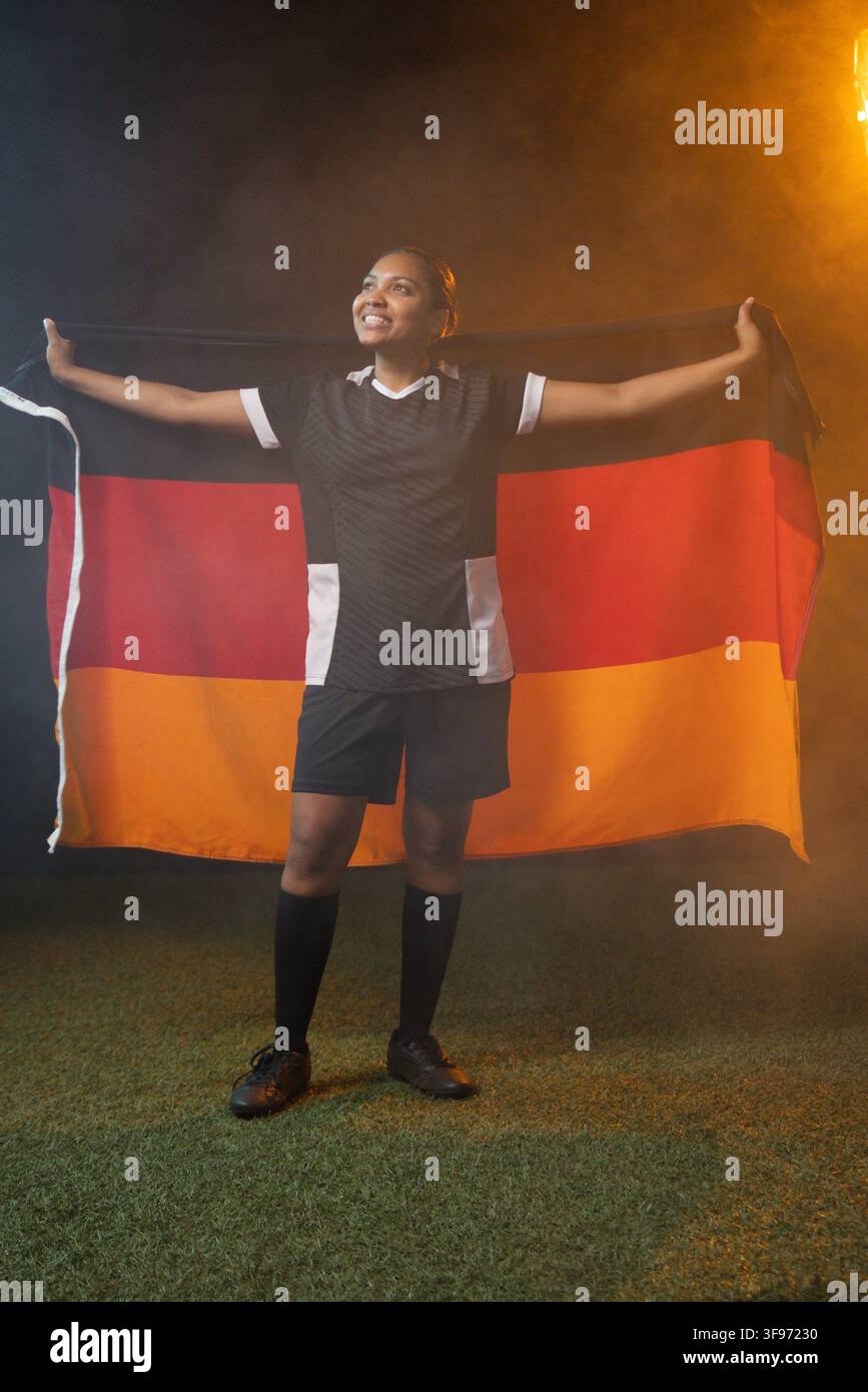 Afrikanische Fußballspielerin hält deutsche Flagge auf Kunstrasen, trägt schwarzes Trikot, Shorts, Fußballschuhe Stockfoto