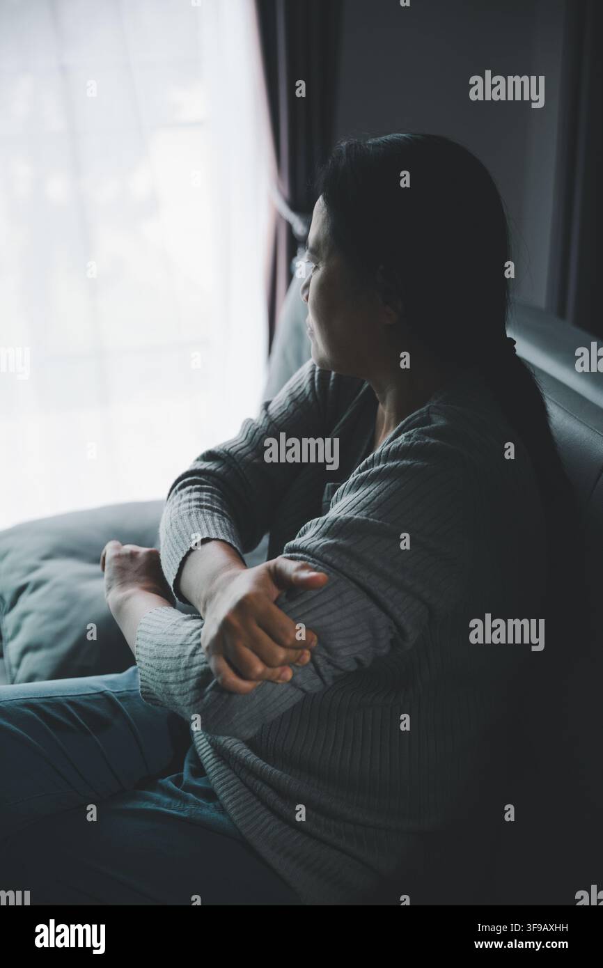 Einsame erwachsene Frau, die sich selbst umarmt, während sie neben einem Fenster in einem dunklen Raum sitzt und Traurigkeit, Angst, emotionale Isolation, Unsicherheit und eine n ausdrückt Stockfoto