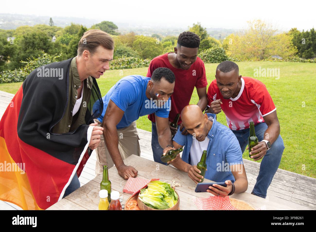 Verschiedene männliche Freunde lehnen sich vor, schauen auf das Smartphone und halten grüne Bierflaschen auf einem erhöhten Deck Stockfoto