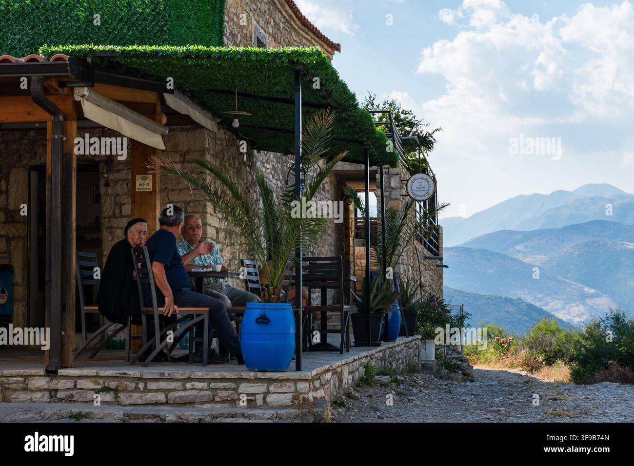 QEPARO FUSHE, ALBANIEN - 28. JULI 2026: Menschen, die in einem kleinen Steinkaffeehaus in den albanischen Bergen sitzen, reale Lebensstil-Reiseillustration mit malerischem Blick a Stockfoto