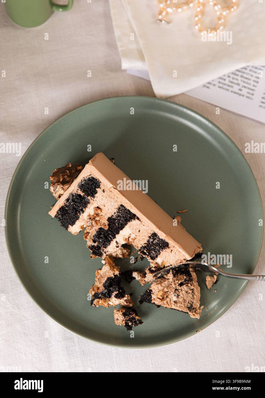 Nahaufnahme einer Scheibe geschichteten Schokoladenkuchens mit cremiger Füllung, serviert auf einem grünen Teller mit einer Gabel und einer Tasse Kaffee im Hintergrund. Stockfoto