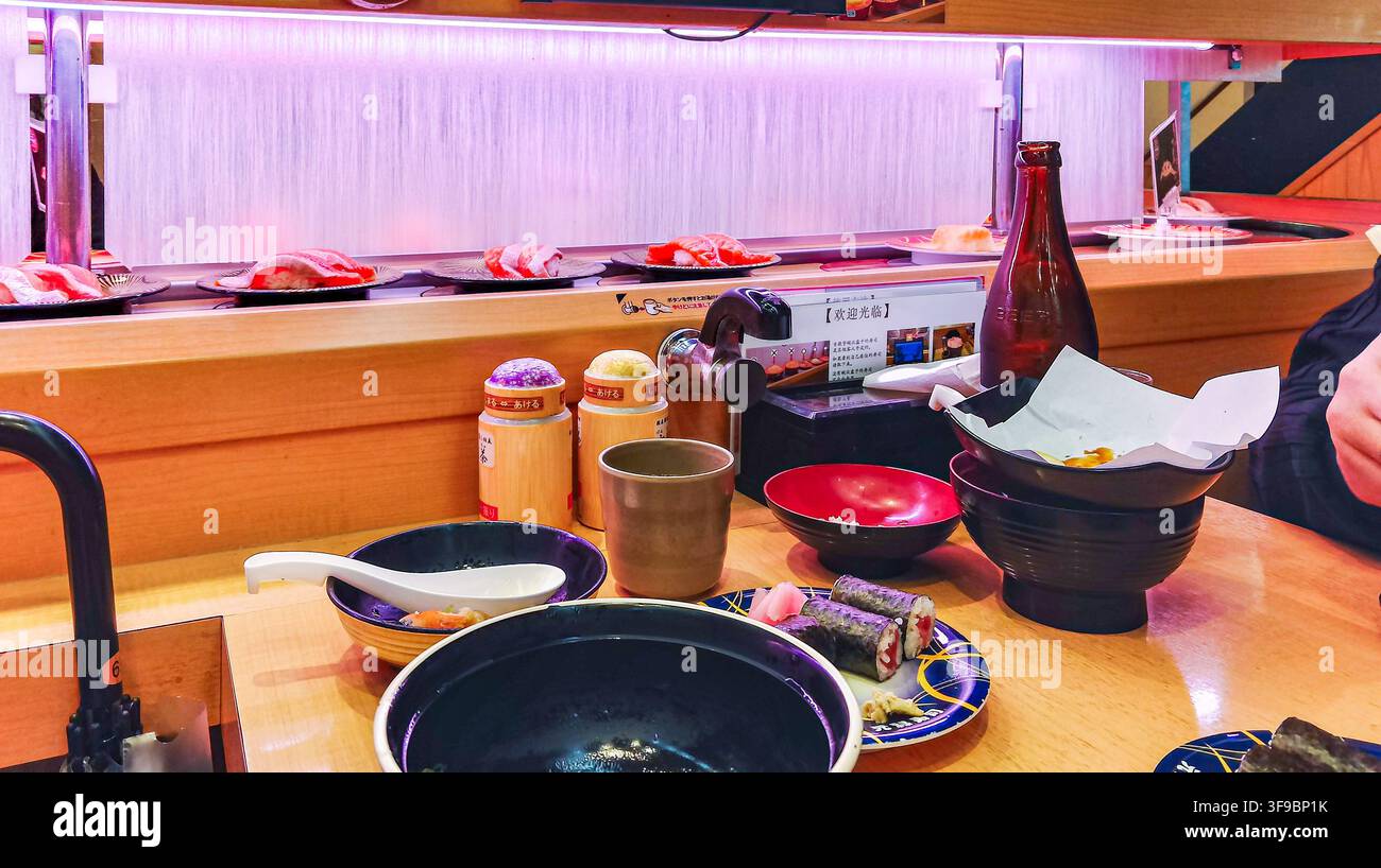 Eine Suppenschale, eine Teetasse und Maki-Rollen auf einer Holzarbeitsplatte mit Sushi-Tellern, die sich auf einem Förderband im Hintergrund eines japanischen Restaurants bewegen. Stockfoto