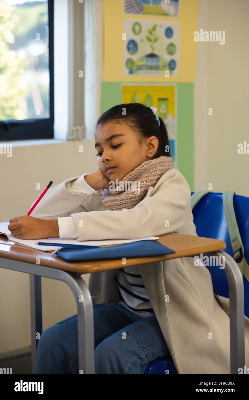Kind afrikanischer Abstammung lehnt sich an den Schreibtisch und schreibt mit einem roten Stift in ein Notizbuch in der Nähe eines blauen Stifteetuis Stockfoto