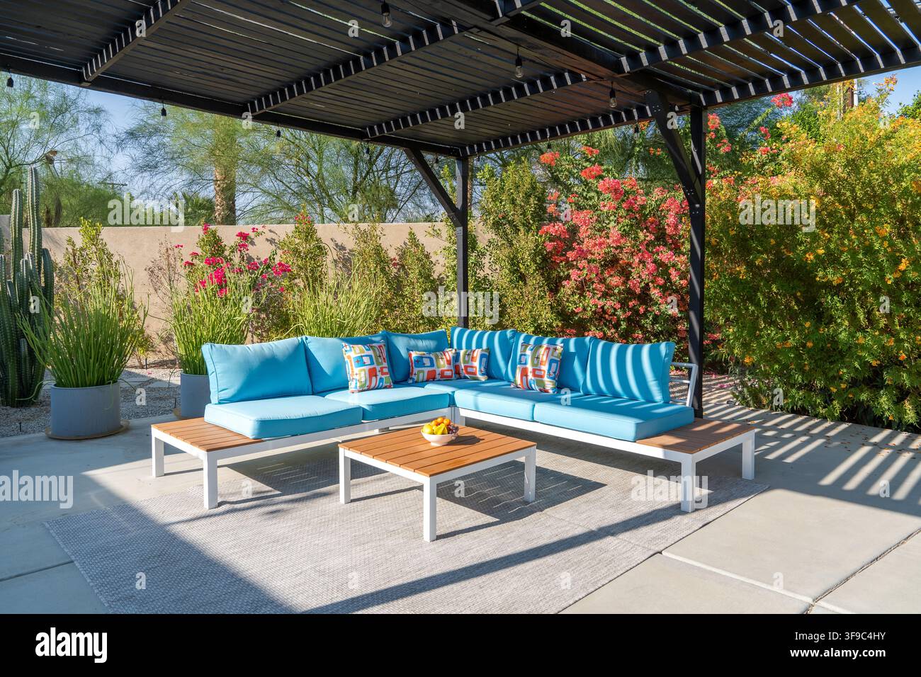 Ein luxuriöser Außenwohnbereich mit Sitzgelegenheiten. Helles Sonnenlicht beleuchtet ein modernes Ecksofa mit blauen Kissen und bunten Kopfkissen unter einer Pergola Stockfoto