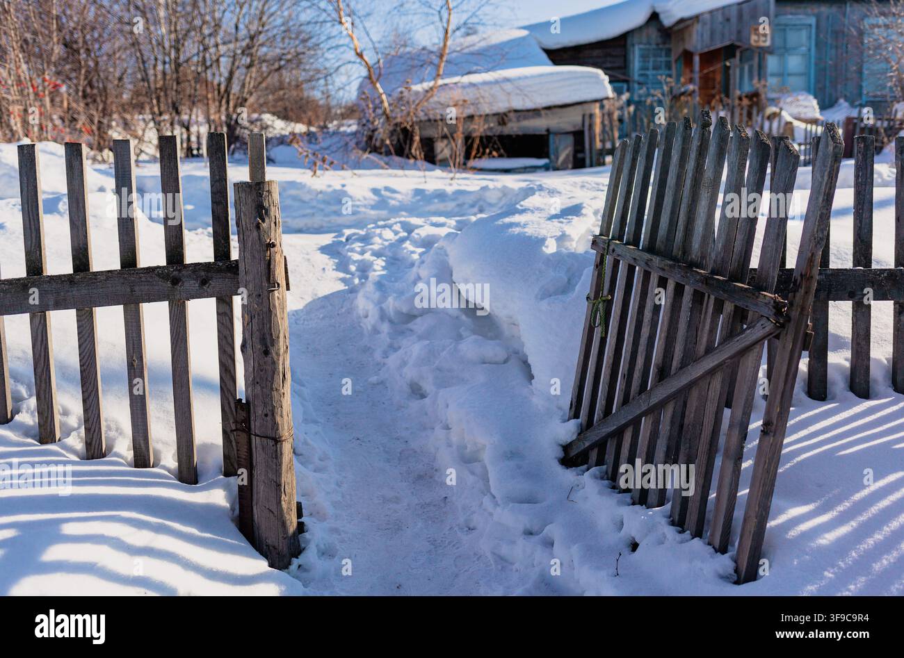 Schneebedeckter Weg durch ein offenes Holztor, der zu einem rustikalen Haus in einer Winterlandschaft mit Bäumen und Schneeverwehungen führt Stockfoto