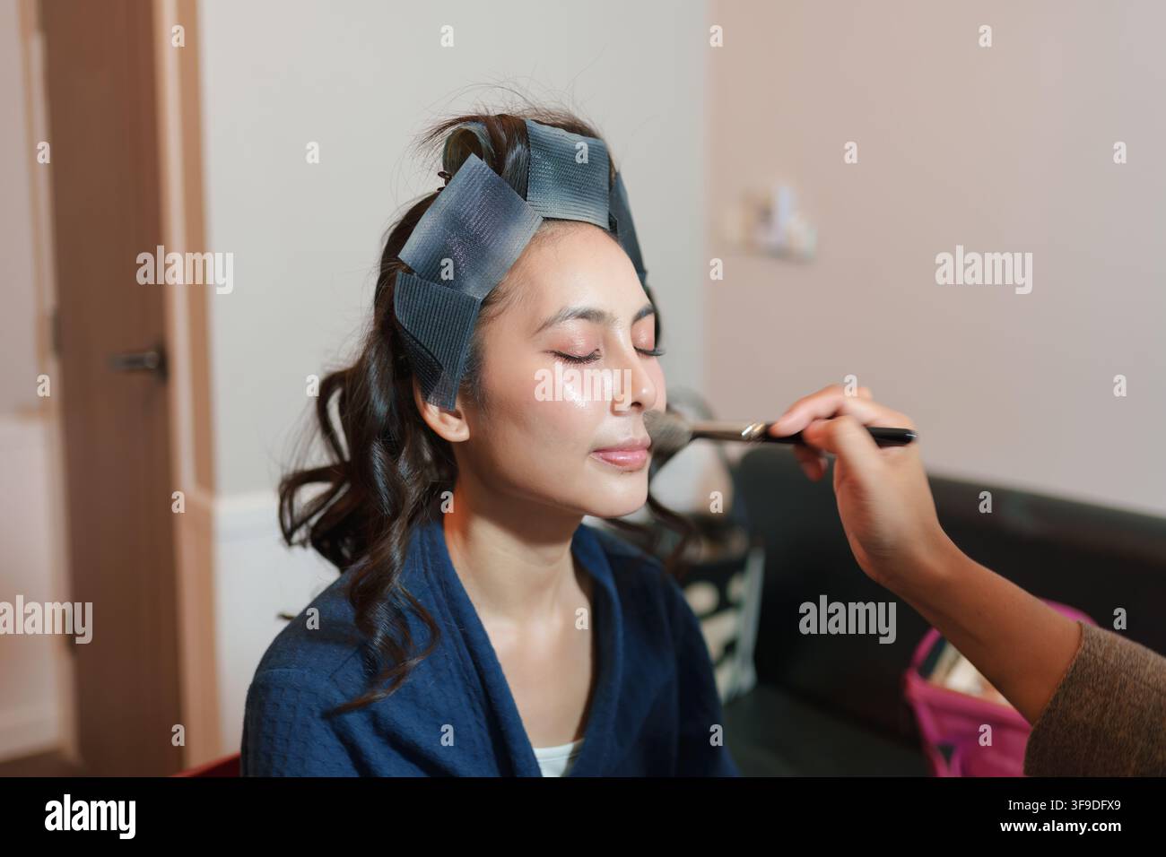 Eine Frau genießt eine sanfte Make-up-Anwendung in einem gemütlichen Innenraum. Das weiche Licht unterstreicht ihre Gesichtszüge und bereichert die Schönheitsroutine. Stockfoto