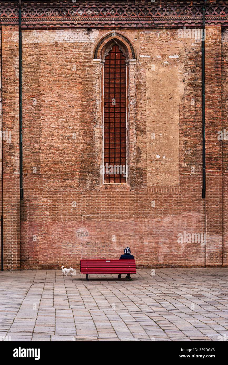 Minimalistische einsame Figur auf roter Bank mit kleinem weißem Hund und roter Backstein-Gotikaußenwand der Basilika im Platz von Venedig Stockfoto