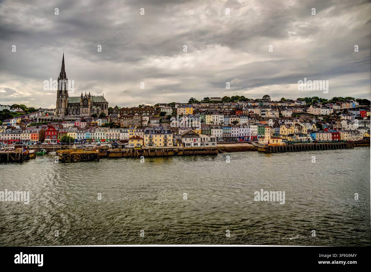 Die St Colman's Kathedrale erhebt sich über dem farbenfrohen Ufer von Cobh unter dramatischen Wolken über dem Cork Harbour. Stockfoto