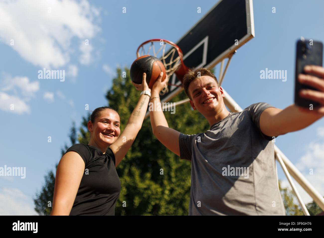 Junge Basketballspieler, die einen erfolgreichen Wurf feiern, während sie an einem sonnigen Tag im Freien in der Nähe eines Basketballplatzes ein Selfie machen Stockfoto