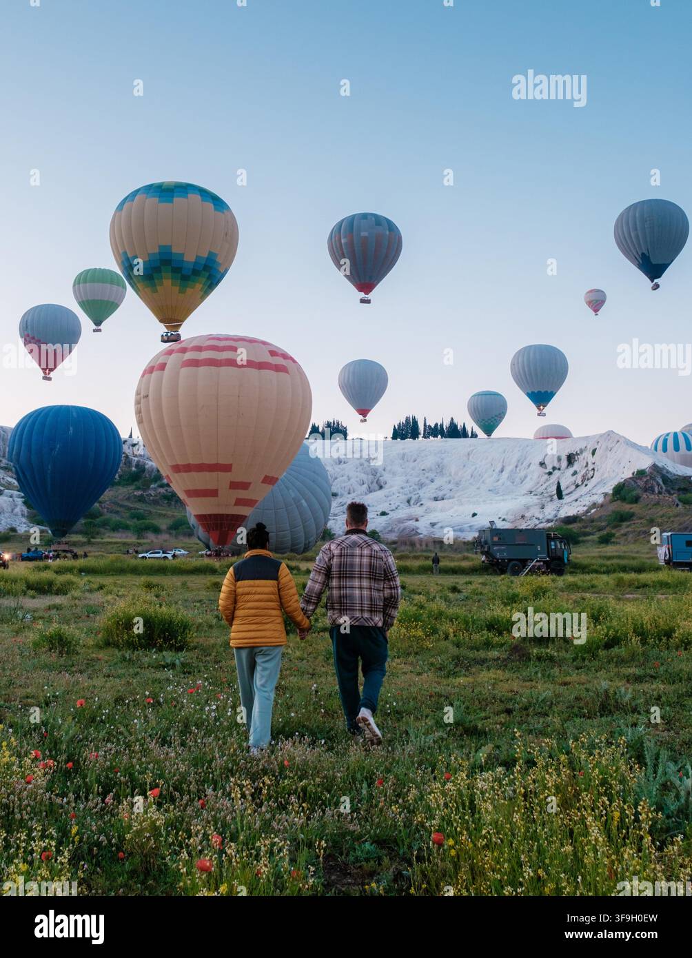 Paar schlendert Hand in Hand durch üppige Felder, während farbenfrohe Heißluftballons den Himmel über Pamukkale, Türkei, füllen. Dieser malerische Ort ist berühmt für seine Travertinterrassen. Stockfoto