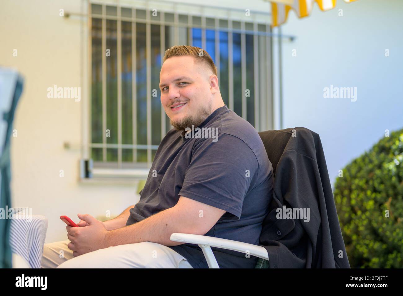 Entspannter junger Mann sitzt draußen auf einer Terrasse, während er ein Smartphone hält und natürlich in die Kamera lächelt. Lässiges Lifestyle-Porträt mit modern Stockfoto