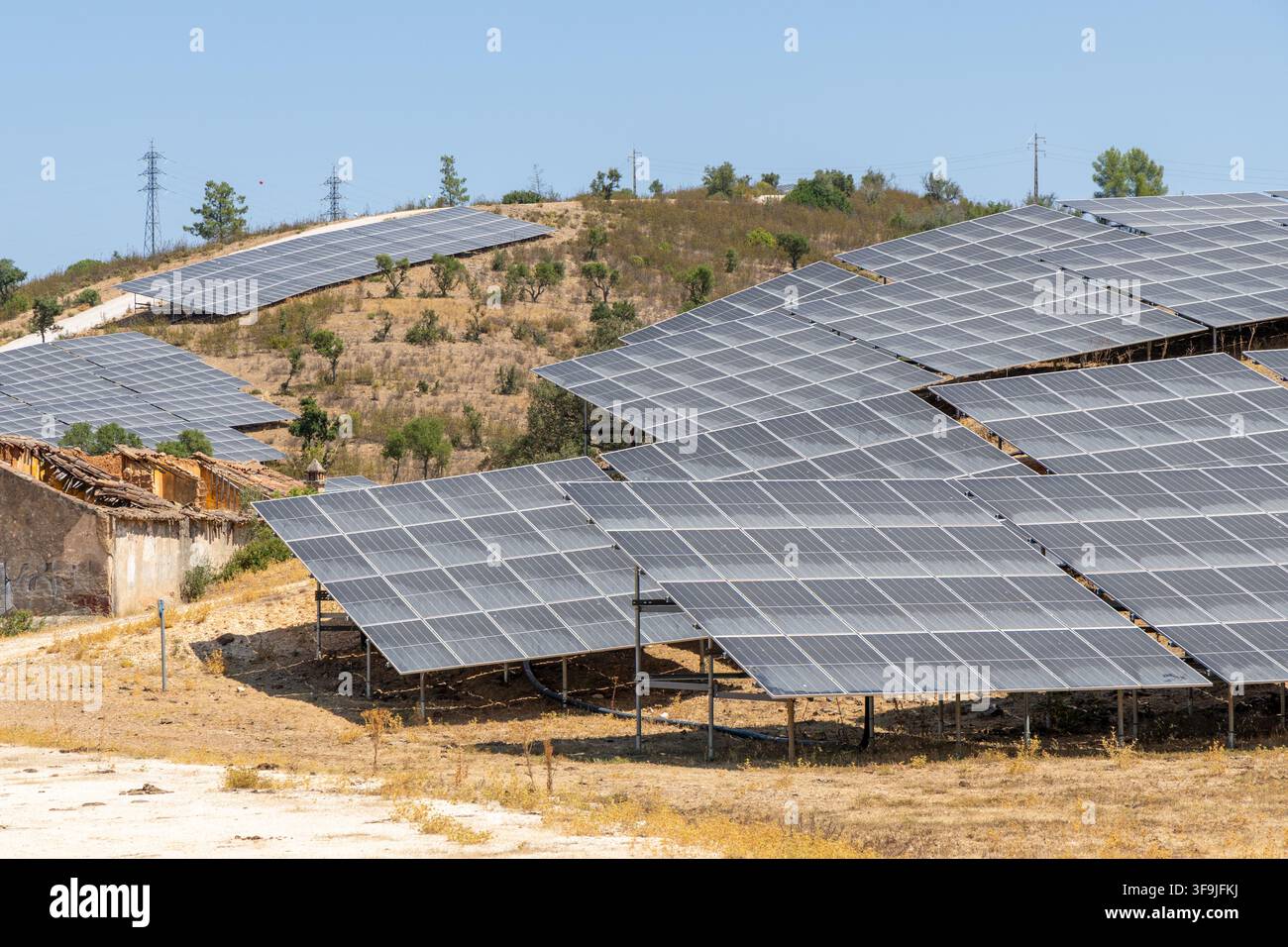 Reihen von Photovoltaikpanelen sind auf einem sonnenverwöhnten Hügel installiert, wobei einige ältere Strukturen in der Nähe sichtbar sind Stockfoto