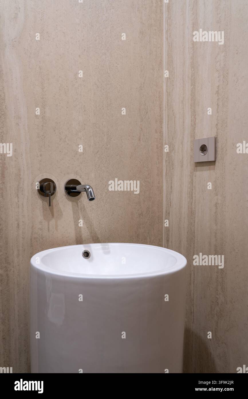 Minimalistisches rundes Standwaschbecken mit Chrom-Wandarmatur vor beiger Travertinwand, keine Personen, modernes Badezimmer Stockfoto