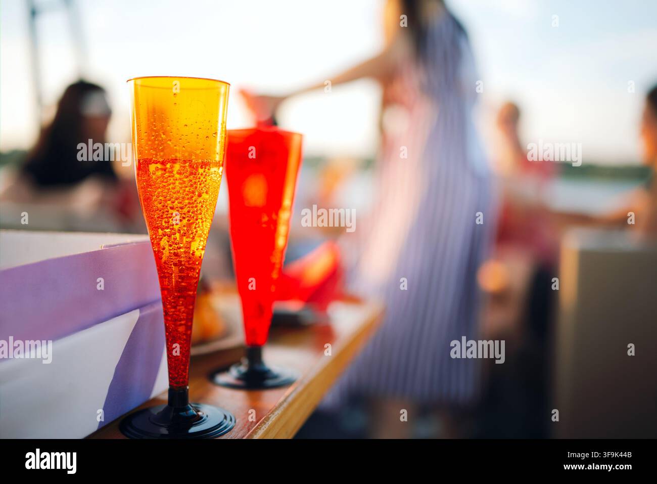 Zwei leuchtend orangefarbene Flöten, gefüllt mit einem sprudelnden Getränk, stehen auf einem Holztisch, mit verschwommenen Figuren von Leuten, die im Hintergrund gesellig sind Stockfoto