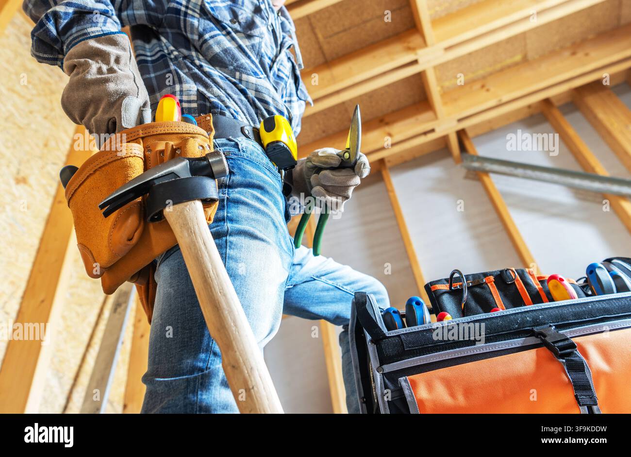 Ein Bauarbeiter steht in einer Werkstatt, trägt Handschuhe und Jeans. Er hält ein Werkzeug, während ein Werkzeuggürtel an seiner Hüfte hängt. Ein Werkzeugkoffer steht offen daneben, Stockfoto