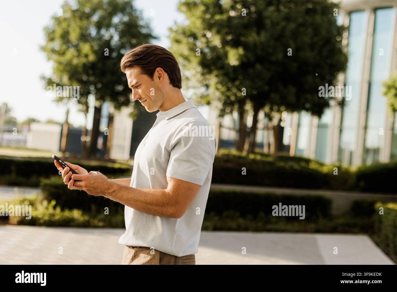 Ein junger Mann steht in einem üppigen grünen Park, vertieft in sein Smartphone, während die Sonne hell über ihm scheint. Stockfoto
