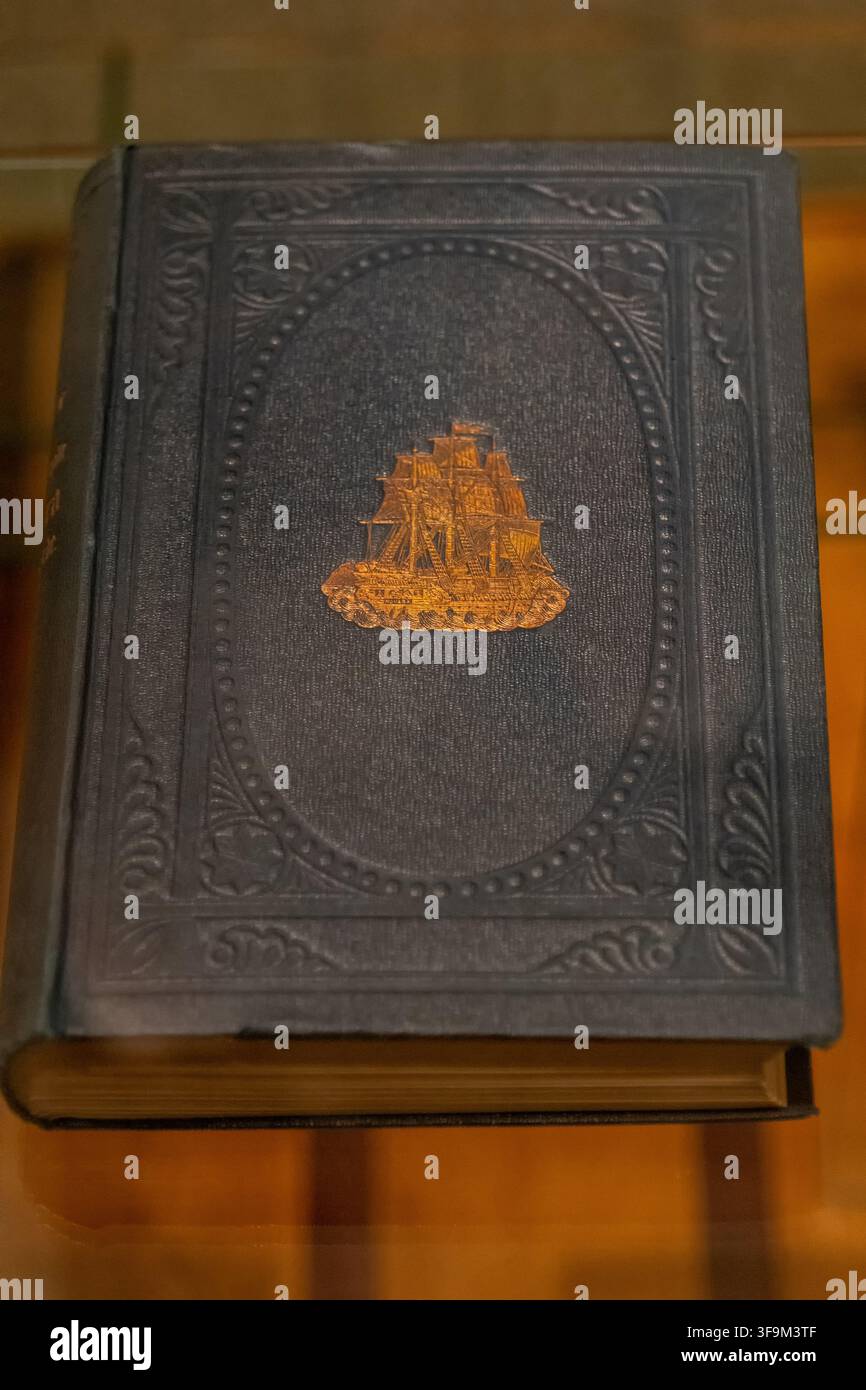 WIEN, ÖSTERREICH, 13. August 2026: Nahaufnahme eines antiken dunklen geprägten Buches mit einem goldenen Segelschiff-Emblem hinter Glas. Stockfoto