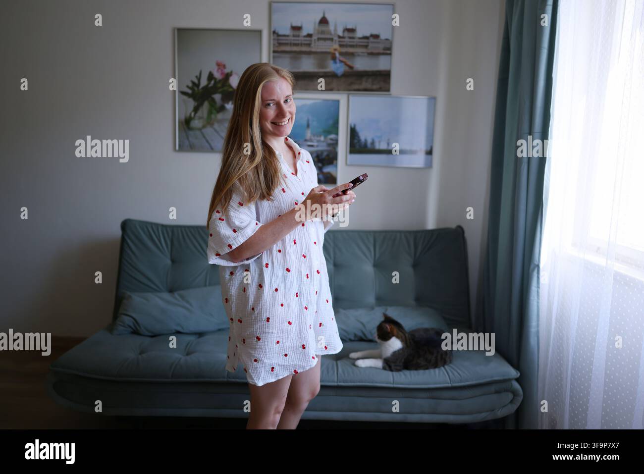 Lächelnde Frau, die auf ihrem Smartphone surft, während sie mit ihrer Katze auf der Couch in einem gemütlichen Wohnzimmer entspannt. Stockfoto
