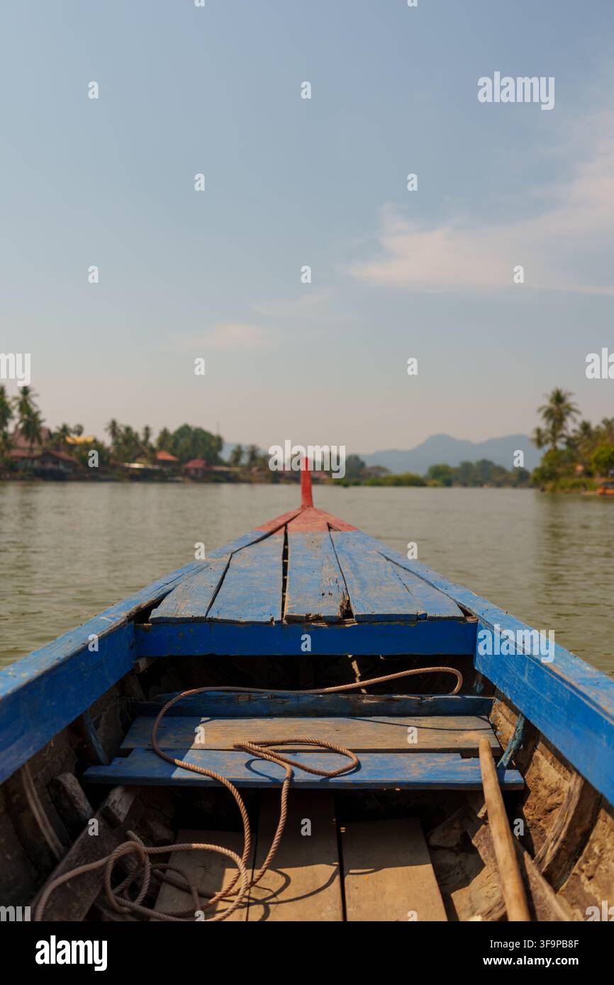 Blick aus einem blauen Holzboot auf dem Mekong, mit einem Seil und einem Paddel im Vordergrund und Häusern am Ufer. Stockfoto