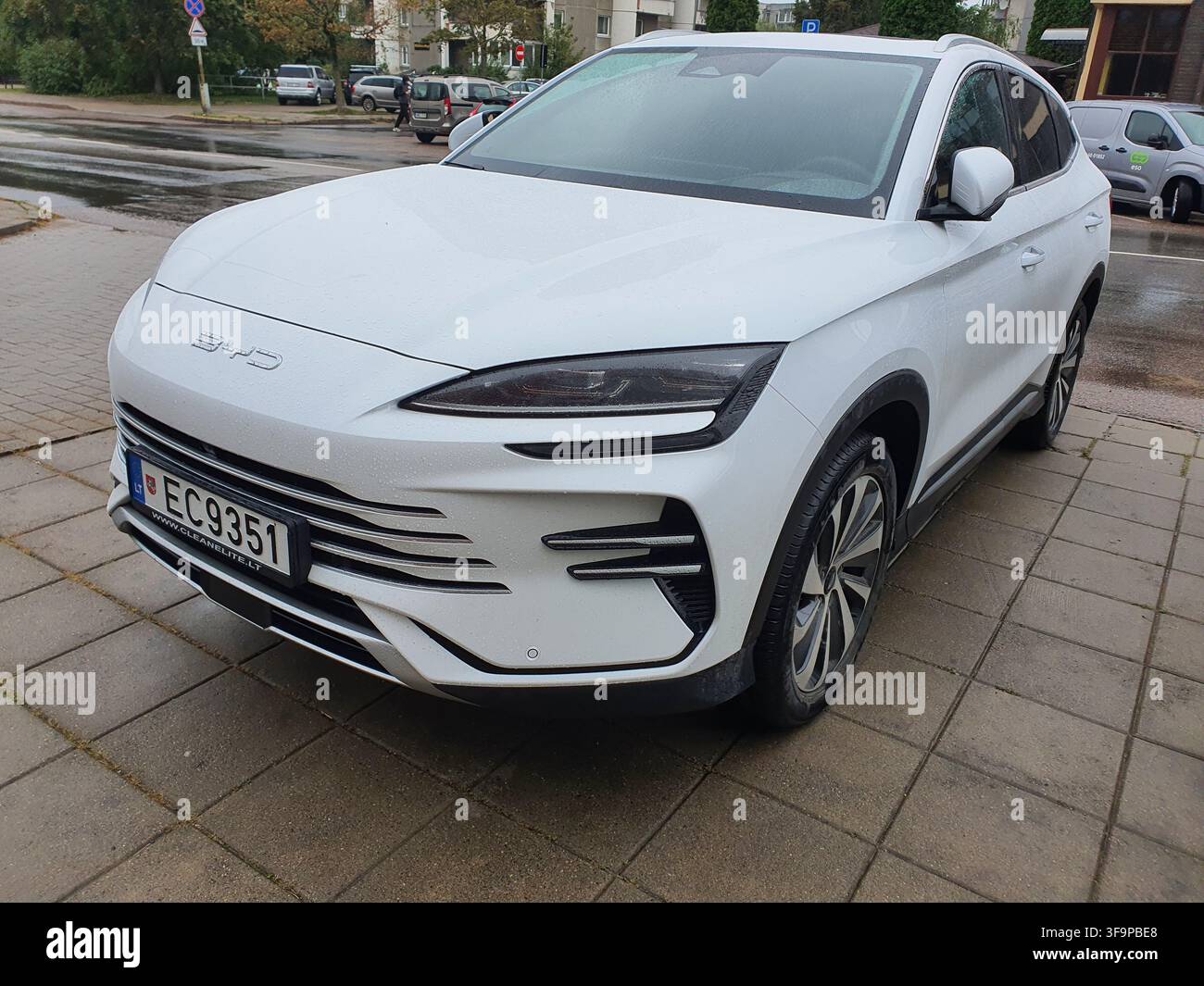 Ein weißer BYD-Elektro-SUV, der auf einem nassen gepflasterten Gehweg in einer städtischen Gegend geparkt ist. Das Fahrzeug zeichnet sich durch ein modernes Design mit schlanken LED-Abblendlichtern aus, eine geschlossene Stockfoto
