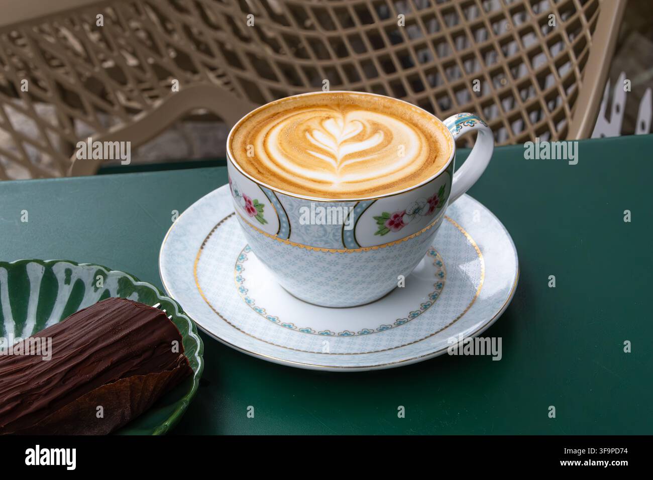 Tasse Cappuccino mit Herz-Schaumkunst auf einer Untertasse neben einem Schokoladengebäck auf einem grünen Cafétisch, der eine gemütliche Kaffeepause schafft. Stockfoto