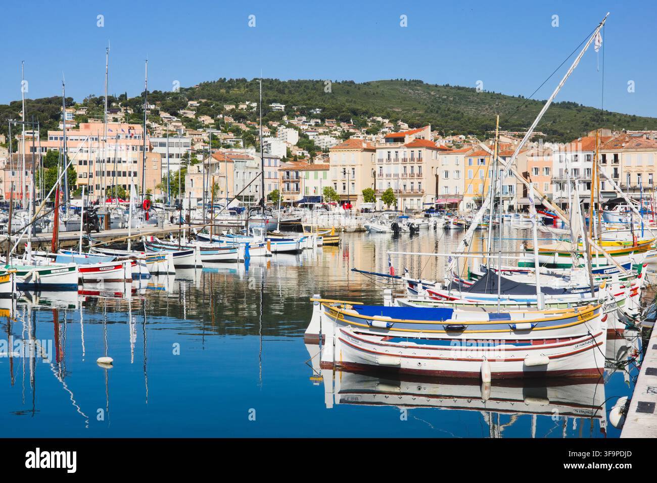 Bunte traditionelle Pointu-Boote, die im alten Hafen von La Ciotat, Frankreich, vor Anker liegen, spiegeln sich in ruhigen Gewässern mit Küstenhügeln im Hintergrund. Stockfoto