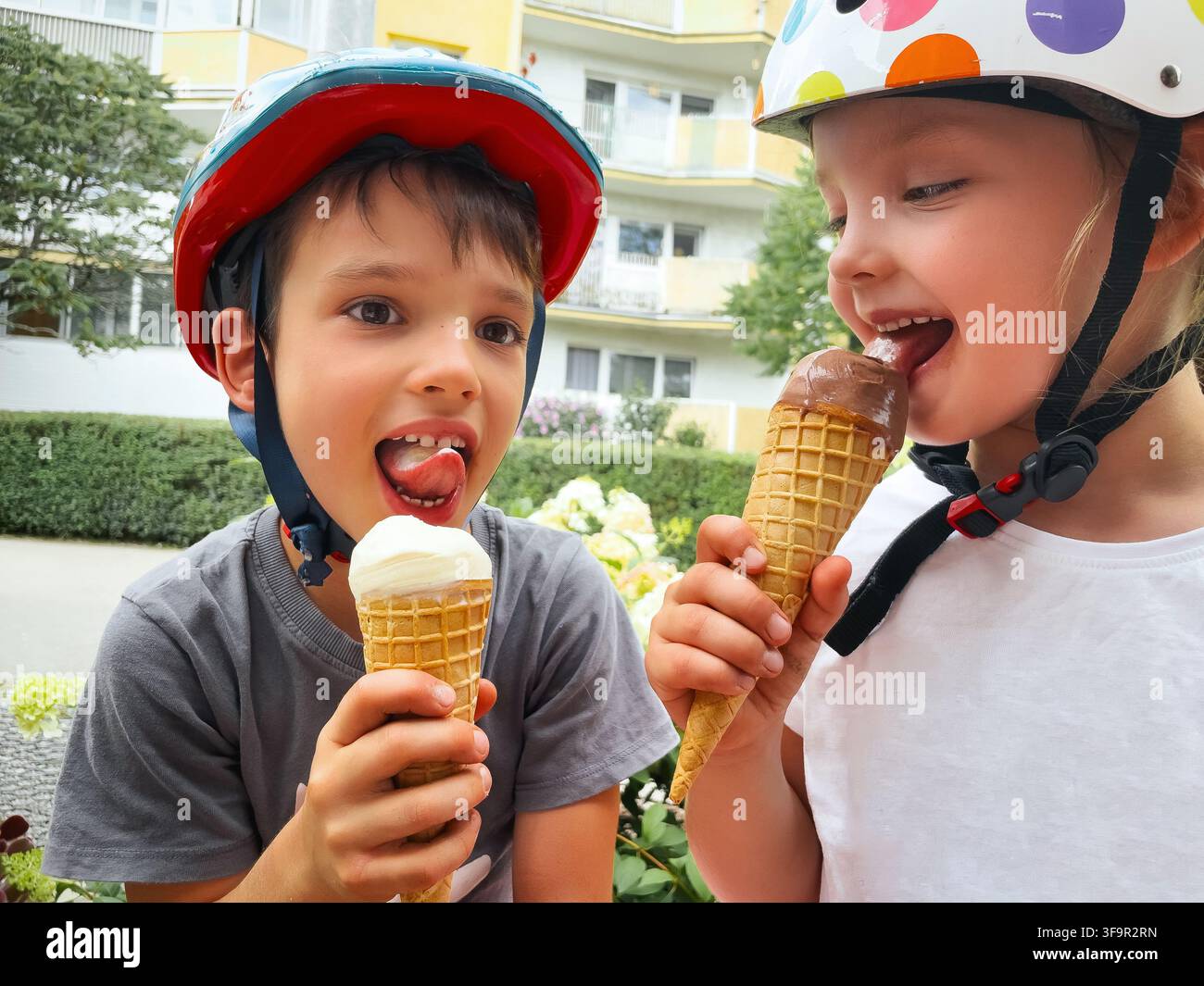 Ein fröhlicher Junge und ein Mädchen, die Helme tragen, genießen Eisbecher draußen. Der Junge leckt sein Vanilleeis, während das Mädchen ihr Schokoladeneis genießt Stockfoto