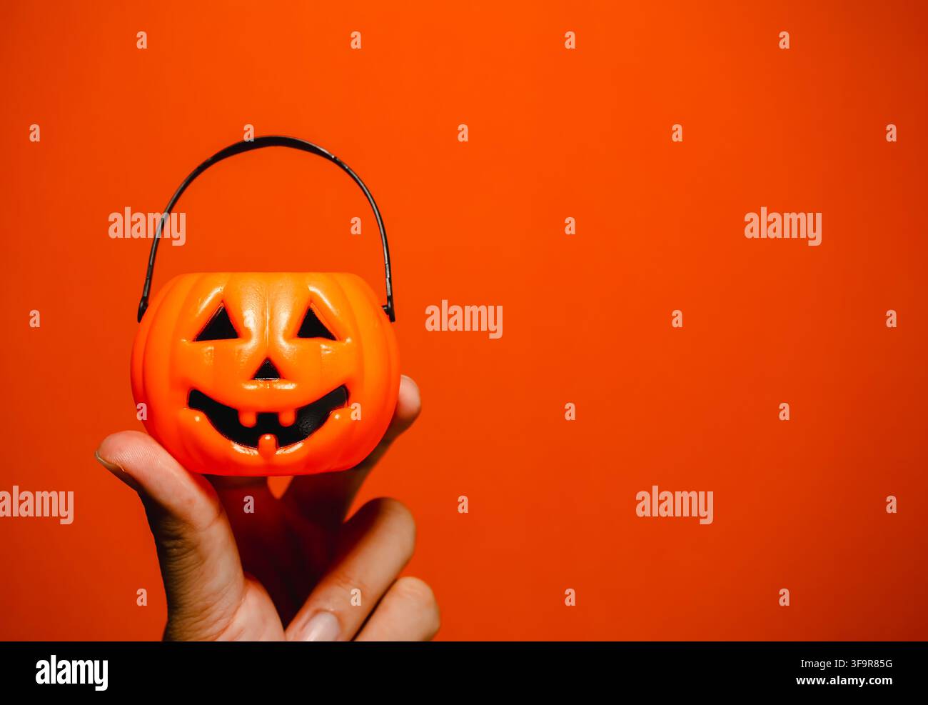 Hand, die einen orangefarbenen Jack-o'-Lantern-Kürbis-Süßigkeitenbecher auf einem lebhaften orangefarbenen Bannerhintergrund mit Kopierraum hält. Halloween-Verkäufe, Party-Promotions, Werbung Stockfoto