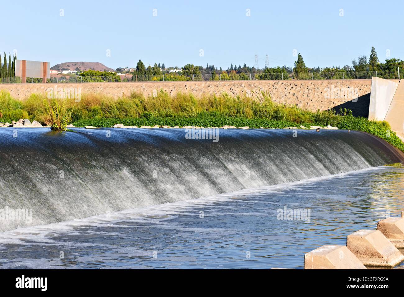 Wasser fließt über einen breiten Betongespeicher am Santa-Ana-Fluss, begrenzt von grüner Vegetation und Hochwasserschutzinfrastruktur in Yorba Linda. Stockfoto