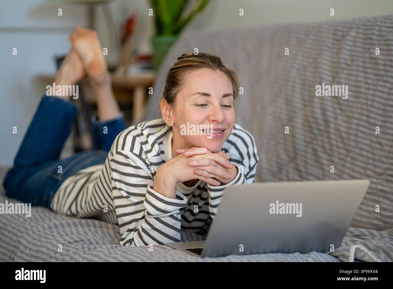 Glückliche entspannte Frau liegt auf dem Sofa zu Hause, ruht sich aus und schaut sich Online-Medieninhalte auf dem Laptop an. Stockfoto