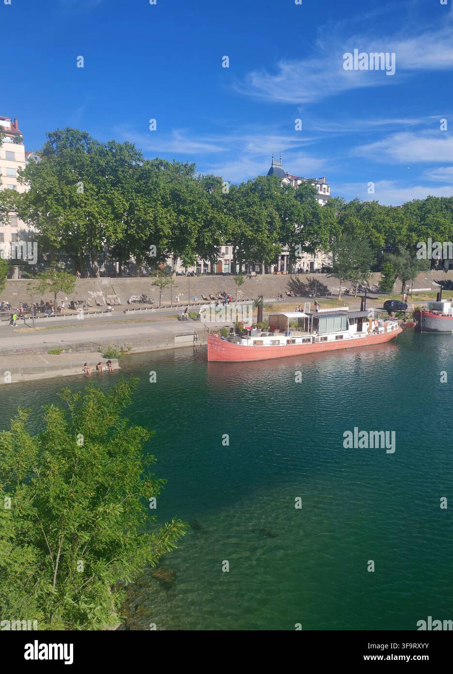 Lyon, Frankreich – 14. Juni 2026: Barge, die auf der Rhône schifft, gesehen von einer Brücke mit kristallklarem blauem Wasser und urbaner Skyline Stockfoto