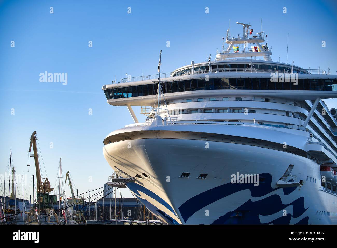Niedrig gewinkelte Aufnahme eines massiven weißen Kreuzfahrtschiffs, das in einem Handelshafen angelegt hat, mit industriellen Kränen im Hintergrund vor einem klaren blauen Himmel du Stockfoto