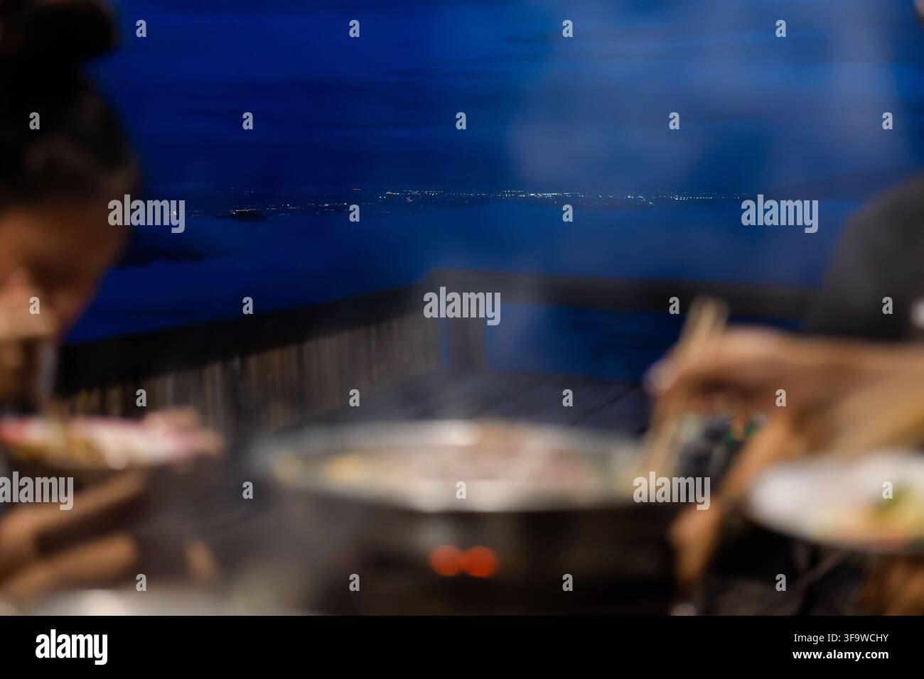 Eine verschwommene Essensszene mit Menschen, die sich an einem Restauranttisch das Essen teilen, warmes Licht, flache Schärfentiefe und blaue Innenraumstimmung, die eine intime Atmosphäre schaffen Stockfoto