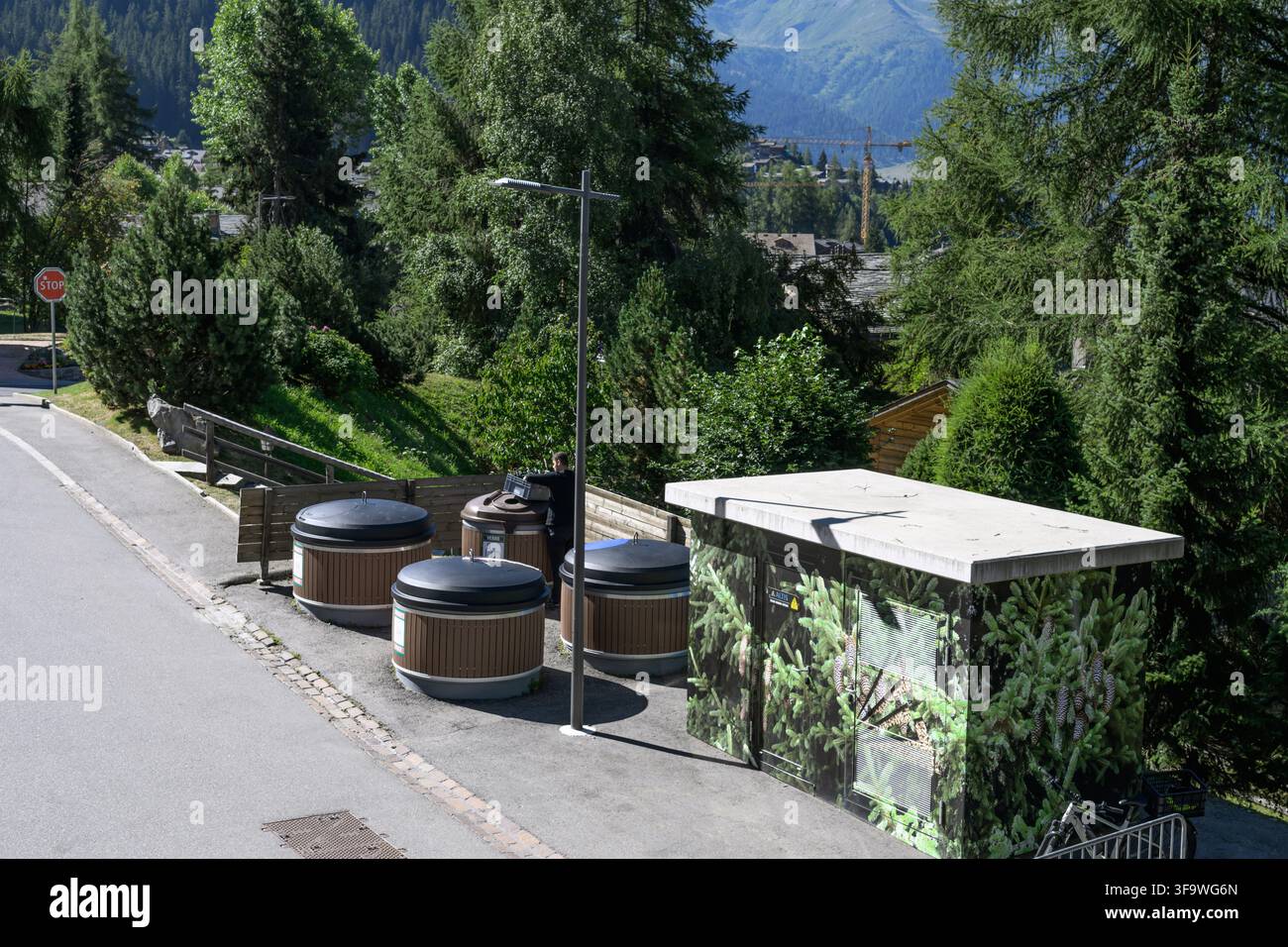 Verbier, Schweiz - 28. Juli 2026: Straßenrandabfälle und Recyclingbehälter neben einer Bergstraße mit Bäumen und alpinen Hängen im Hintergrund. Stockfoto