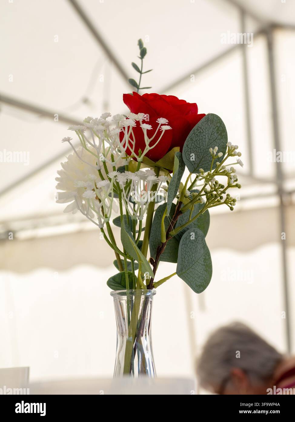 Eine rote Rose und weiße Blumen, die in einer Glasvase drinnen unter einer Zeltstruktur arrangiert sind. Stockfoto
