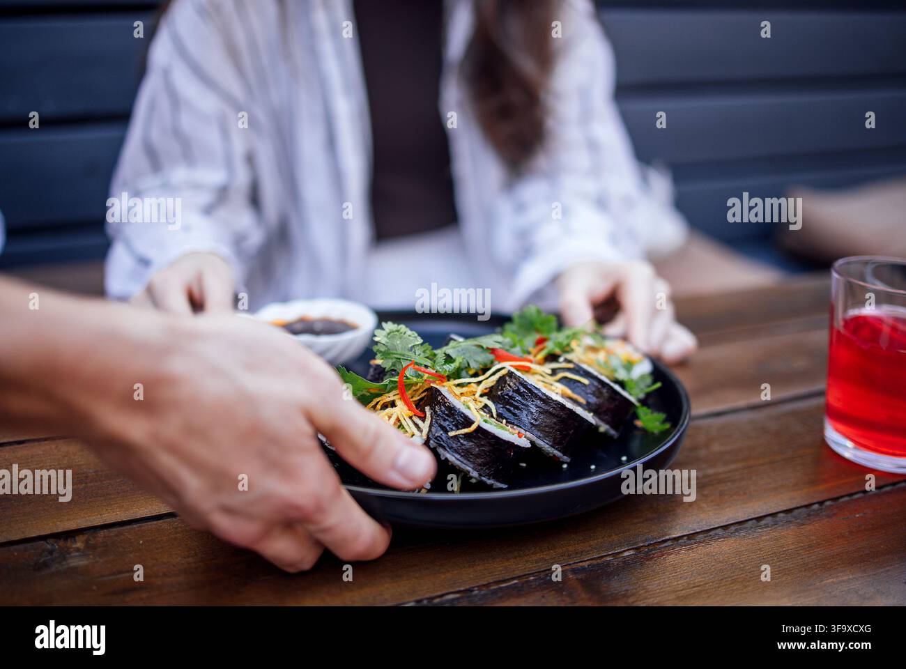 Eine Hand präsentiert einen Teller mit Sushi-Rollen und Garnierungen einer Frau, die an einem Holztisch sitzt, wobei ein Getränk und eine Dip-Sauce in der Nähe sichtbar sind Stockfoto