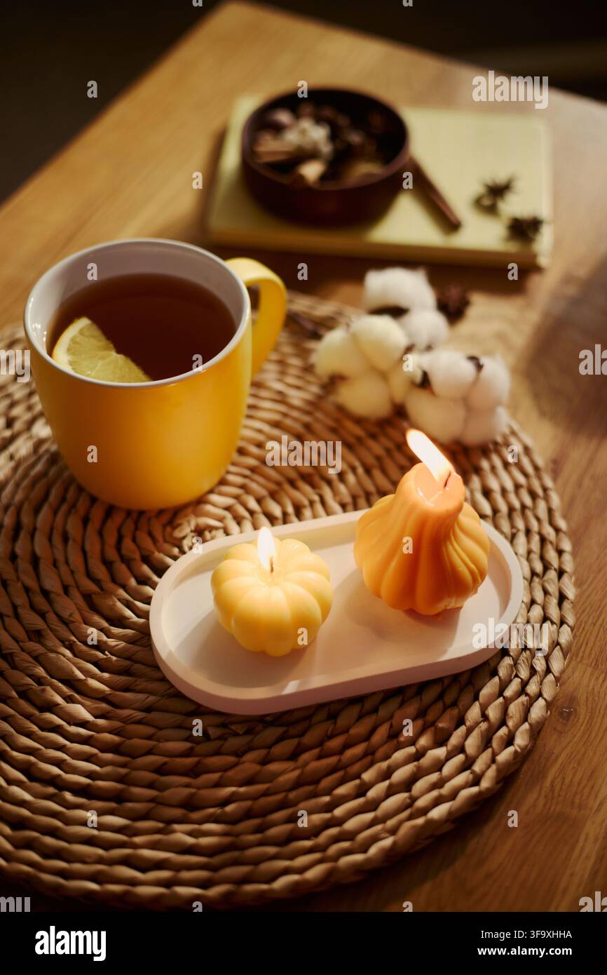 Vertikales Bild von brennenden, kürbisförmigen Kerzen und einer Tasse heißem Tee mit Zitrone auf einer geflochtenen Platzdecke, das Herbst-Wellness, gemütliche Wohnraumdekoration und Selbstpflege-Marketingkampagnen fördert Stockfoto